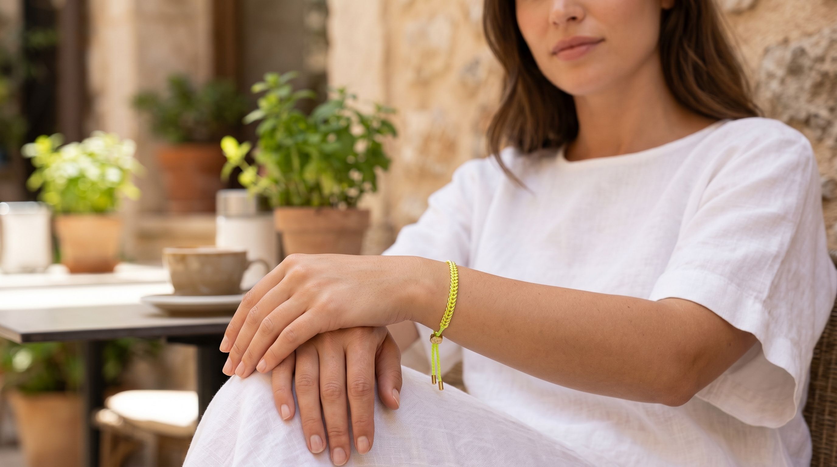Female model wearing Iztac neon yellow and gold woven bracelet on her wrist, resting her hand on her lap, outdoors with a coffee cup in a bright cafe setting.