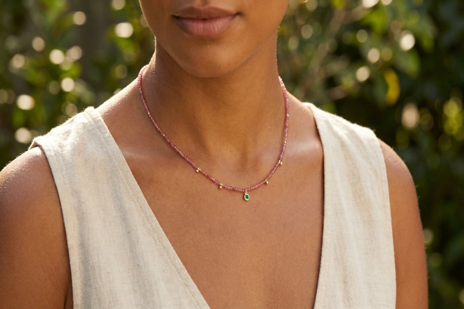 Close-up of female model in linen dress wearing Jasmine gold and pink tourmaline gemstone beaded necklace, showing delicate chain, shimmering pink beads, and tiny golden charms against outdoor greenery.
