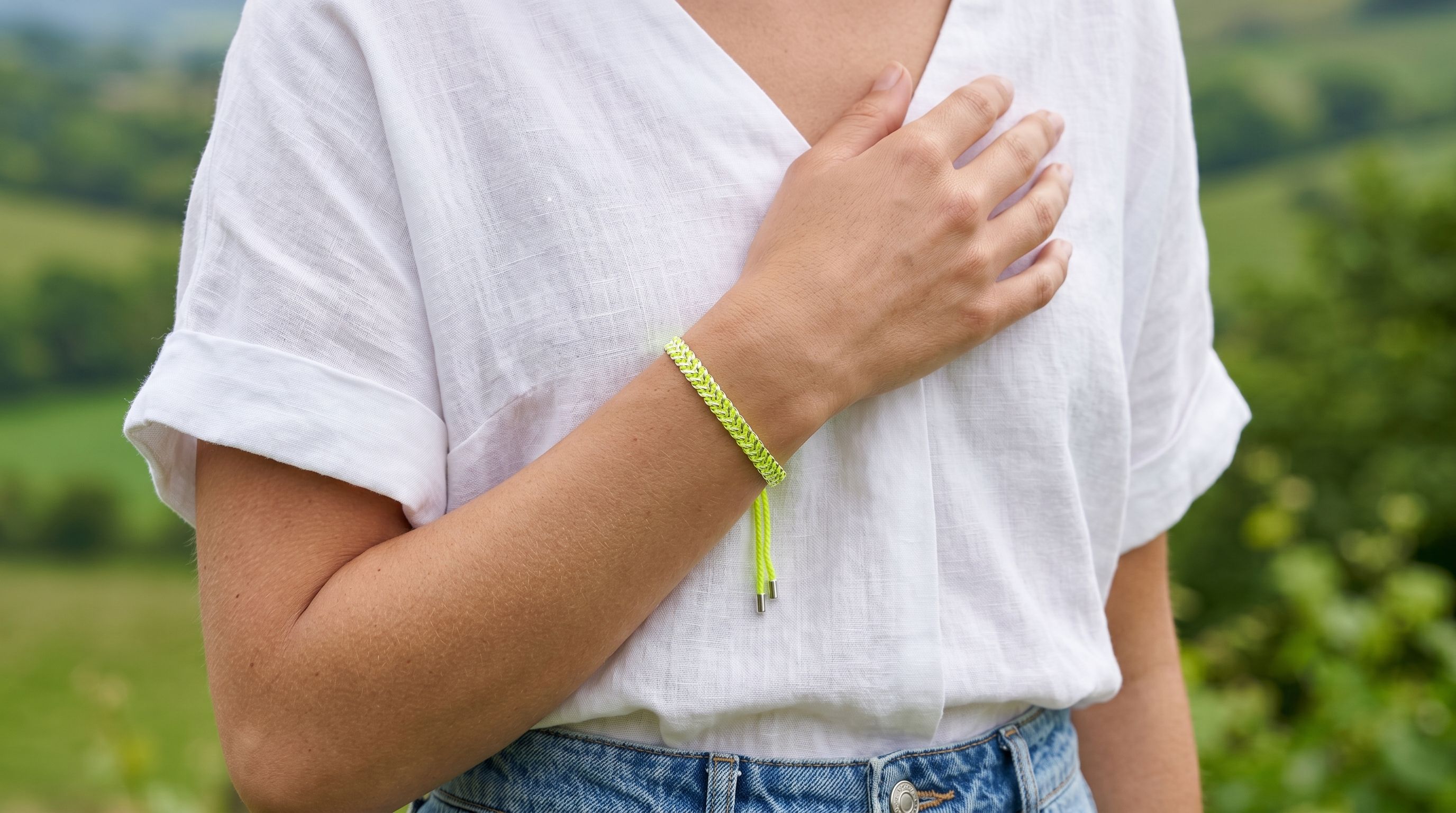 Feminine model wearing neon yellow and silver woven bracelet in lush green outdoor setting, showcasing detailed bracelet design and vibrant colors.