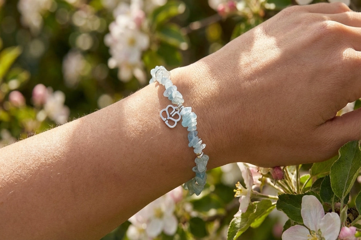 Close-up of a female model's wrist wearing a silver stretch beaded bracelet with aquamarine crystals and a silver charm, set in a spring garden.